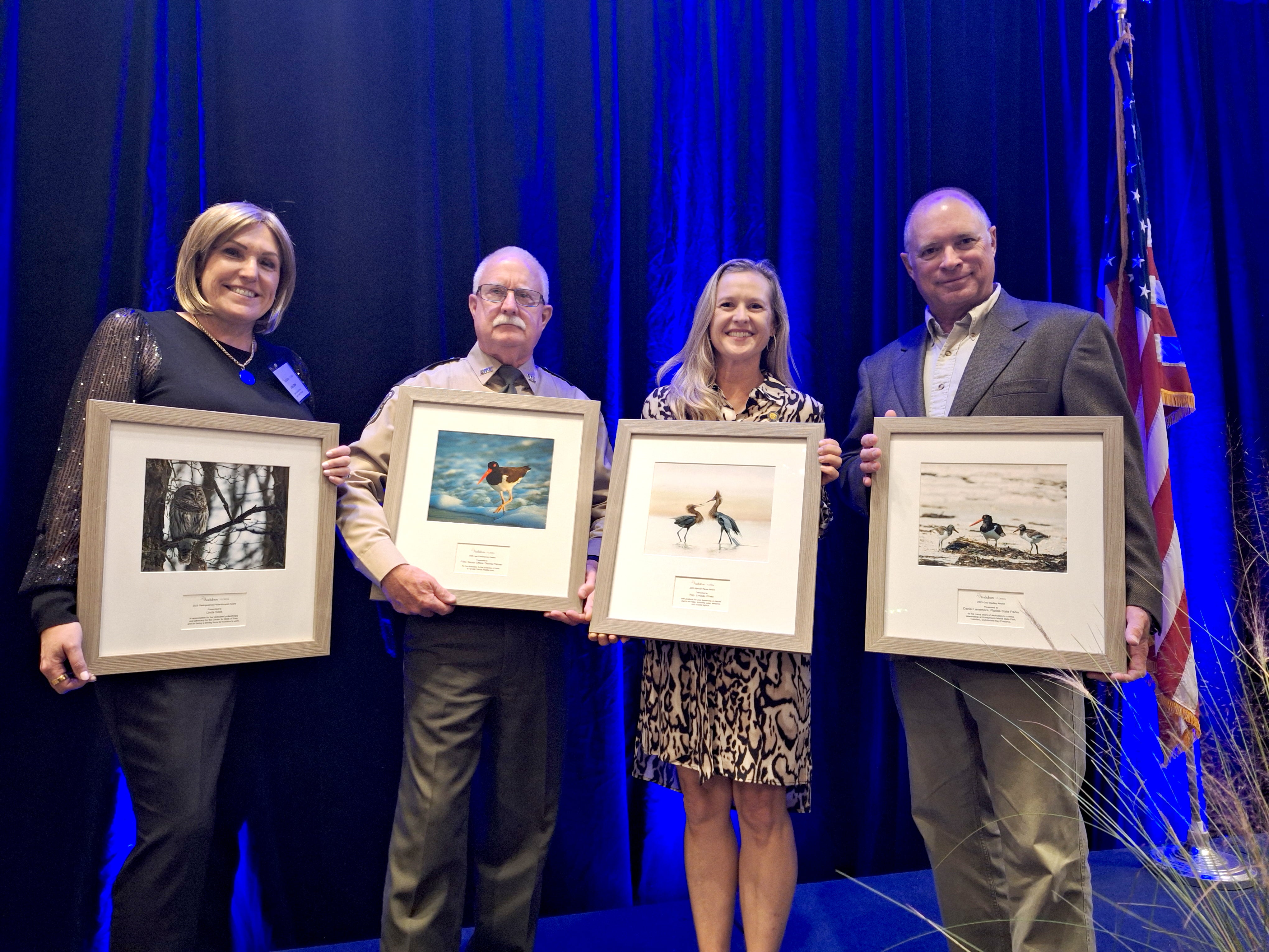 Four people on a stage, each holding a framed photograph